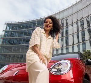 Frau steht lächelnd vor einem roten GWM ORA 03 und berührt ihr Haar; im Hintergrund ist die Glasfassade eines modernen Gebäudes zu sehen.