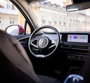 Moderner Innenraum eines GWM ORA 03 mit Blick auf das Lenkrad und ein breites digitales Display; durch die Fenster sind mehrstöckige Gebäude in einer Stadt zu sehen.