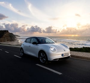 Weißer KGWM ORA 03 fährt bei Sonnenuntergang auf einer Küstenstraße; links ragen Klippen auf, rechts ist das Meer mit Wellen und farbigem Abendhimmel zu sehen.